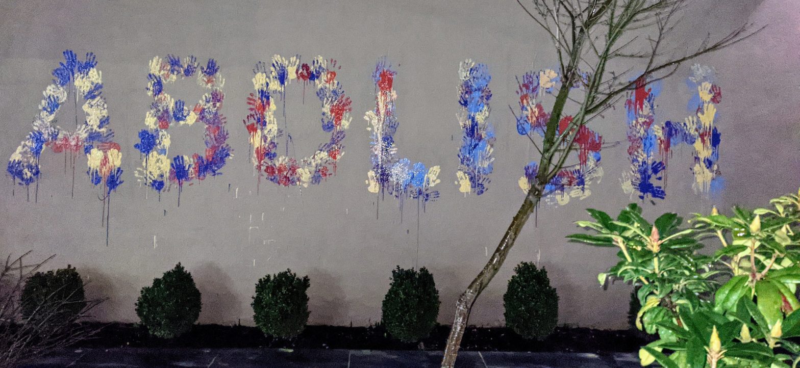The word "abolish" in large capital letters on a wall by blue, red, and yellow paint handprints